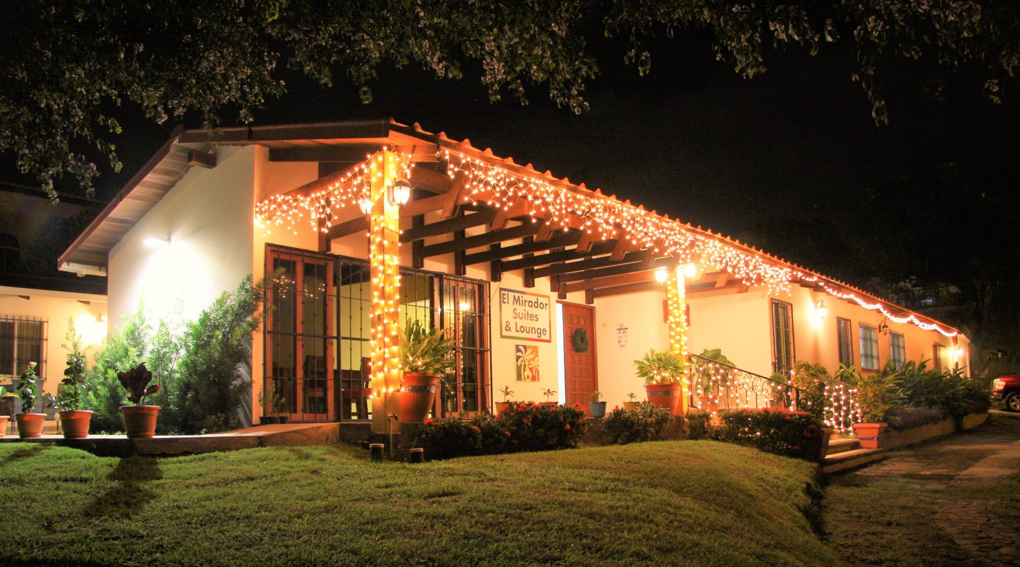 El Mirador Suites and Lounge at night: the white house with a tiled roof, string lights along the eaves, a lit porch and the hotel sign by the door