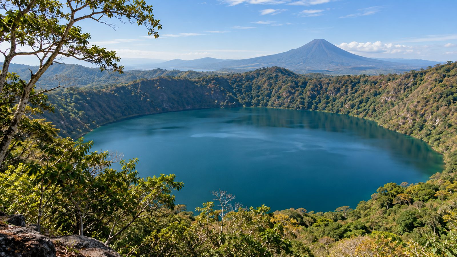 A volcanic crater lagoon ringed by dry tropical forest, with a cone-shaped volcano on the horizon