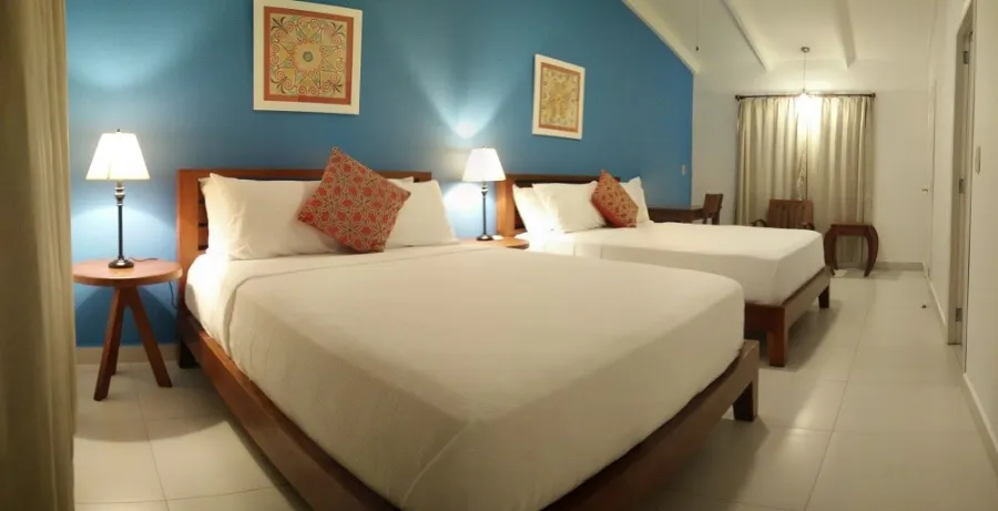 A room with two beds, a blue wall, framed folk art and bedside lamps