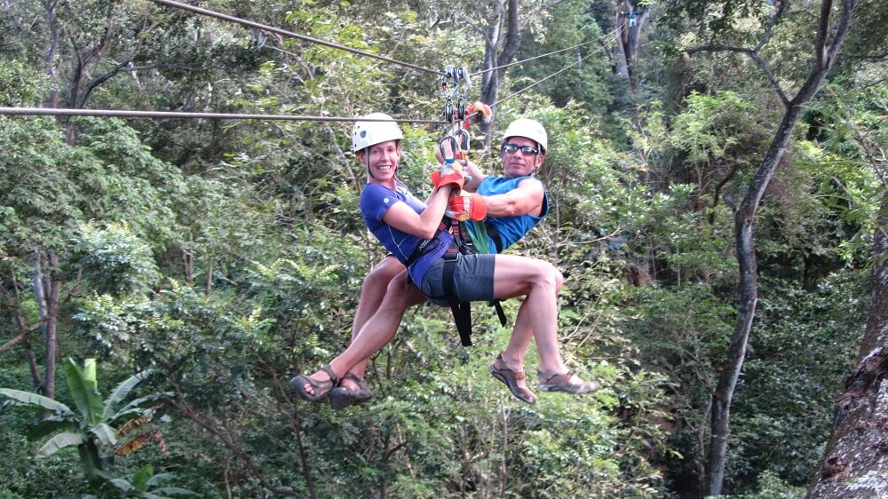 Two guests in helmets and harnesses riding a zip-line together over the forest canopy
